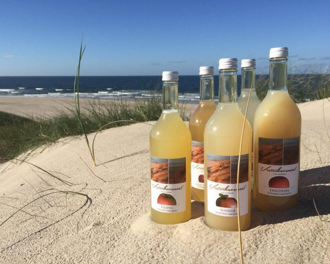 Smagfulde æblemoste fra Danske Vesterhavsmost sat i sandet på stranden. Find dem i Hobby Haven